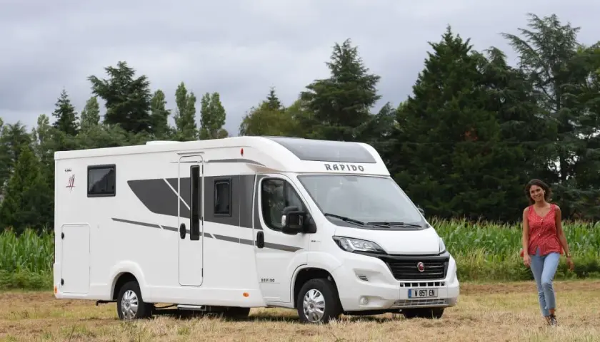 Un camping-car Rapido C86 blanc, moderne, garé dans un champ. Une jeune femme souriante se tient à droite. Avis Rapido C86.