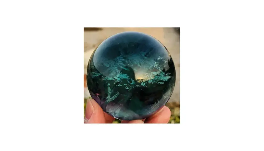 Hand hält eine polierte Kugel aus Regenbogenfluorit, die in tiefen Blau- und Grüntönen schimmert. Perfekt, um Regenbogenfluorit kaufen.