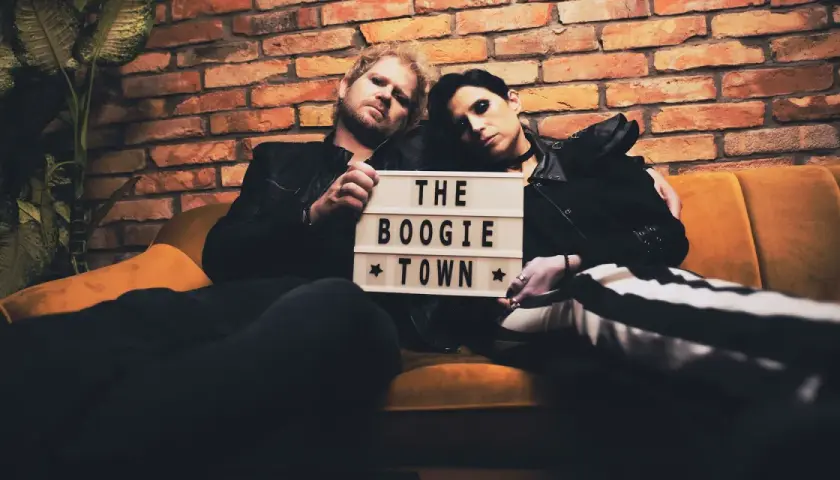 Para siedzi na kanapie, trzymając tabliczkę z napisem "THE BOOGIE TOWN".
