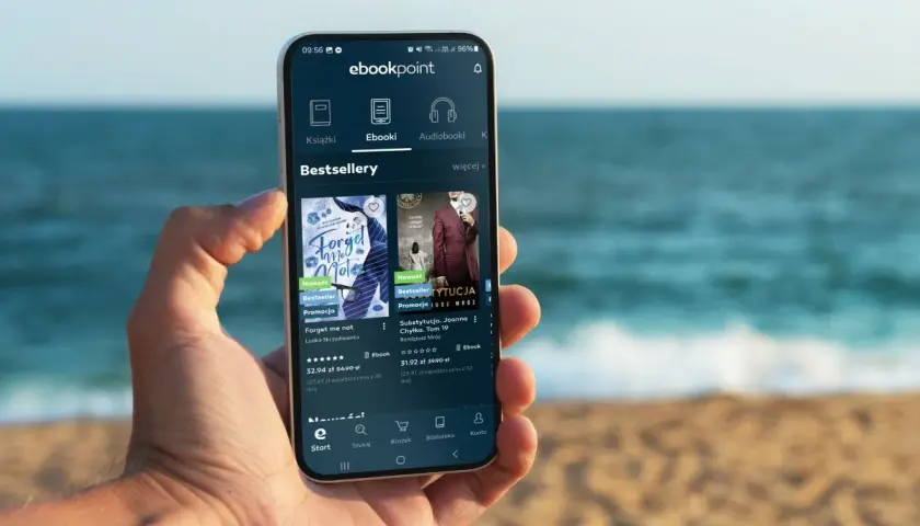 Na smartfonie w dłoni wyświetla się aplikacja ebookpoint z bestsellerami. Idealny poradnik przetrwania na wakacjach, by nie nudzić się na plaży.