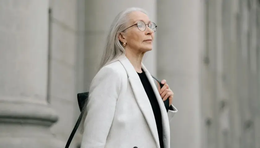 Donna con lunghi capelli grigi, occhiali e un cappotto bianco elegante, con uno sfondo architettonico in tono grigio.
