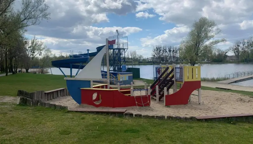 Kolorowy plac zabaw w kształcie statku na piaszczystej plaży nad jeziorem Średzkim. W tle widać zjeżdżalnie wodne i spokojną taflę wody.