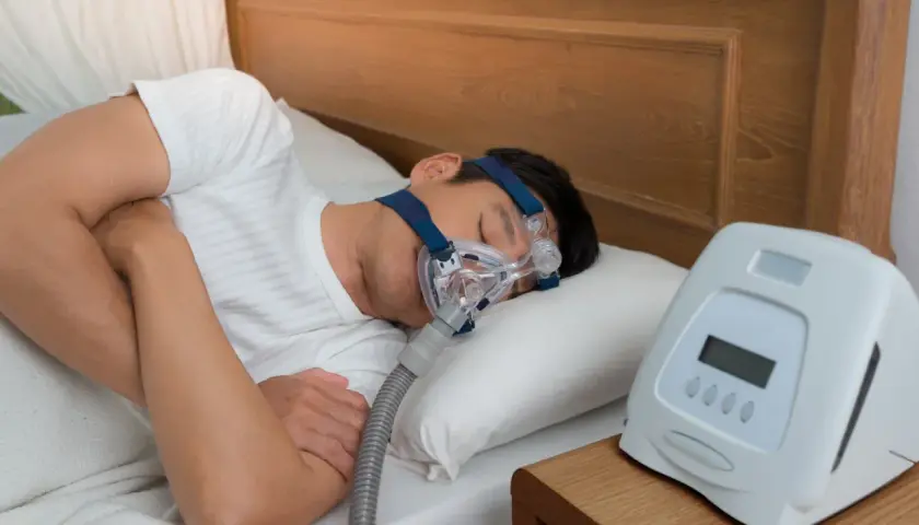 Mann schläft mit CPAP-Maske, um zu wenig Sauerstoff im Schlaf Symptome zu behandeln. Ein Gerät steht daneben.