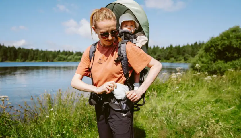 Frau mit kraxe baby am Seeufer, umgeben von grüner Natur und Bäumen.
