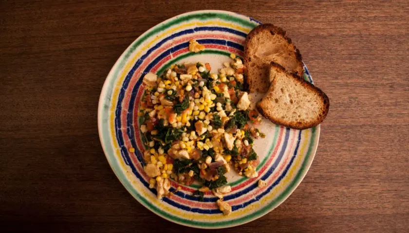 Revuelto de tofu con maíz y espinacas, servido con pan tostado en un plato colorido.