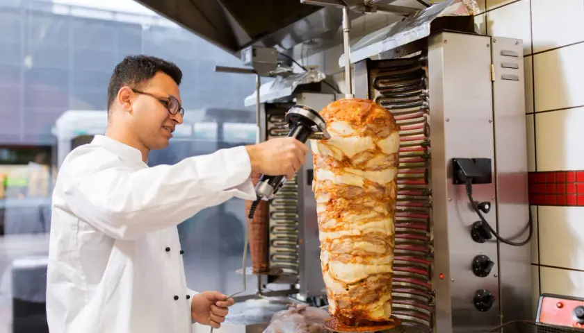 Kucharz w białej kurtce kroi mięso z pionowego rożna na kebab.