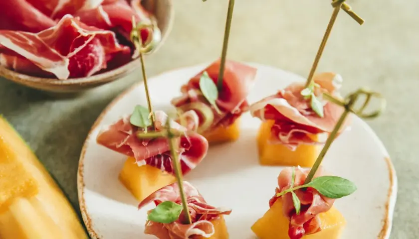 Une présentation originale de melon et jambon, des brochettes savoureuses sur une assiette blanche, avec un melon entier sur le côté.