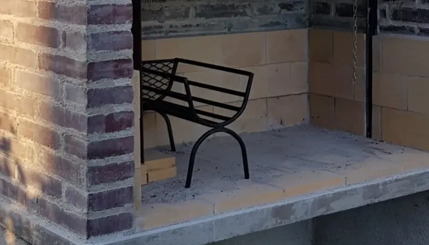 En delvis färdigbyggd grill av tegelstenar. En svart metallbänk står inne i utrymmet.