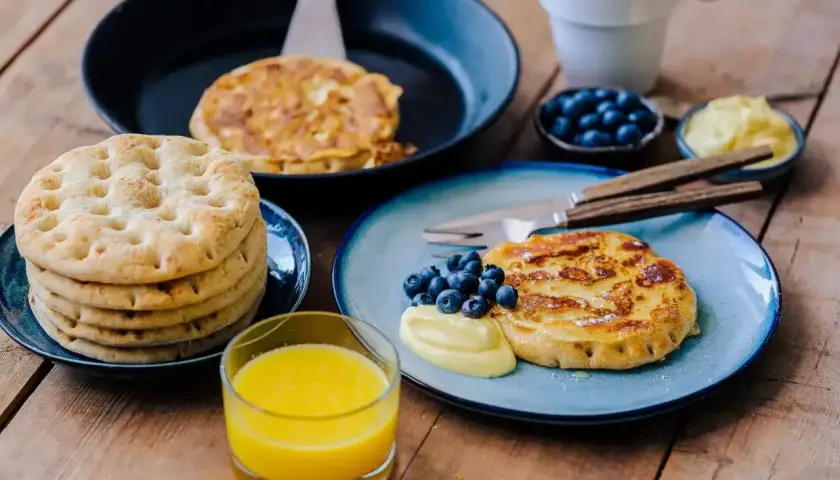 Fattiga riddare med blåbär och vaniljsås på en träyta. En stapel tunna pannkakor och ett glas apelsinjuice.