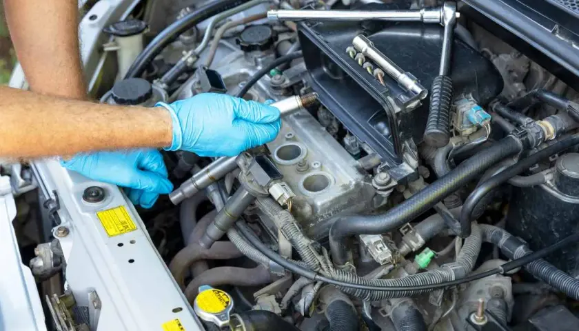 Mano con guante azul sosteniendo una bujía, lista para el cambio de bujías en el motor de un coche. Herramientas a la vista.