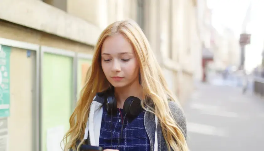 Dziewczyna z blond włosami i słuchawkami na szyi przegląda telefon, zastanawiając się, jak zacząć rozmowę z chłopakiem.