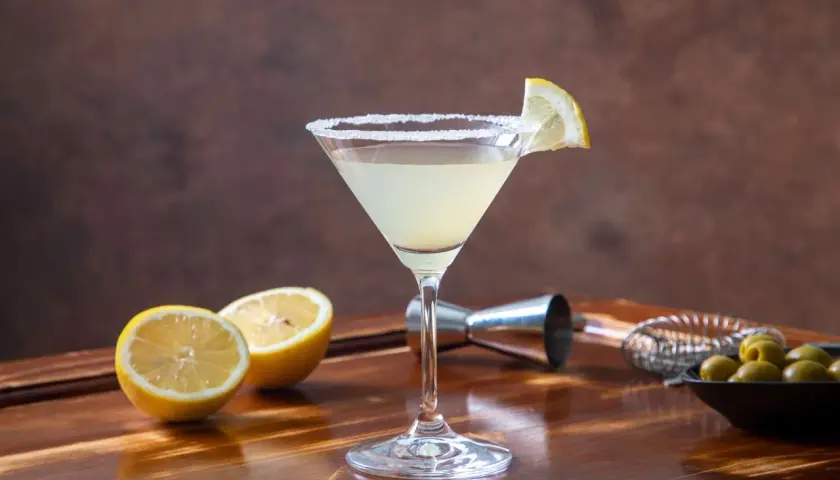 Elegancki martini bianco drink z solnym rantem, ozdobiony plasterkiem cytryny. Obok leżą połówki cytryn i miseczka oliwek.