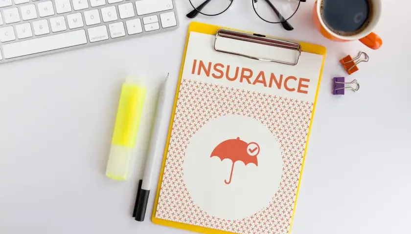 Ubezpieczenie zdrowotne bez pracy: dokument z napisem "INSURANCE" i ikoną parasola. Obok klawiatura, okulary, kawa.