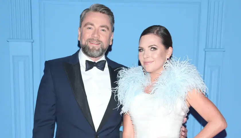 Mąż Oli Kwaśniewskiej, Piotr, w eleganckim garniturze u boku żony w białej sukni z piórami.