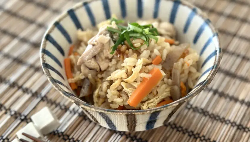 A delicious takikomi gohan recipe served in a striped bowl, featuring rice, chicken, carrots, and green onions.