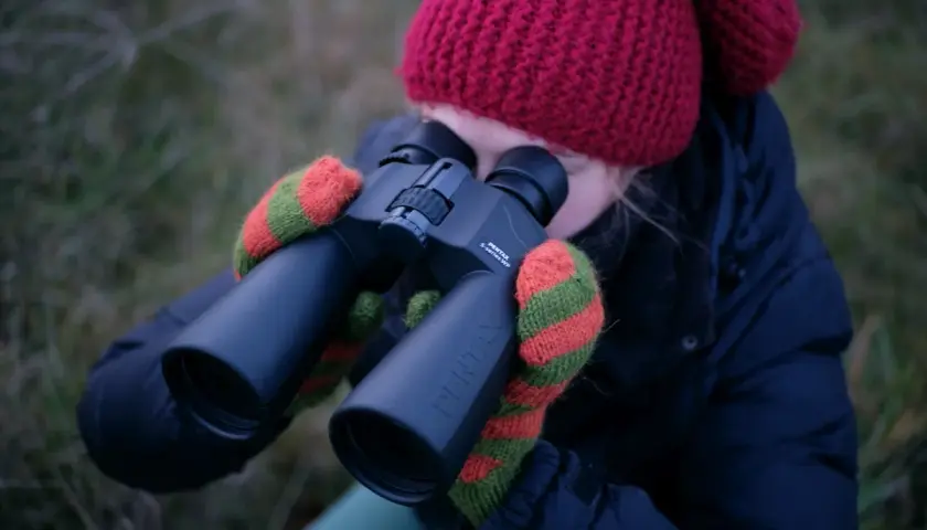 Dziewczyna w czerwonej czapce i rękawiczkach obserwuje niebo przez lornetkę Pentax. Idealna lornetka do obserwacji nieba.