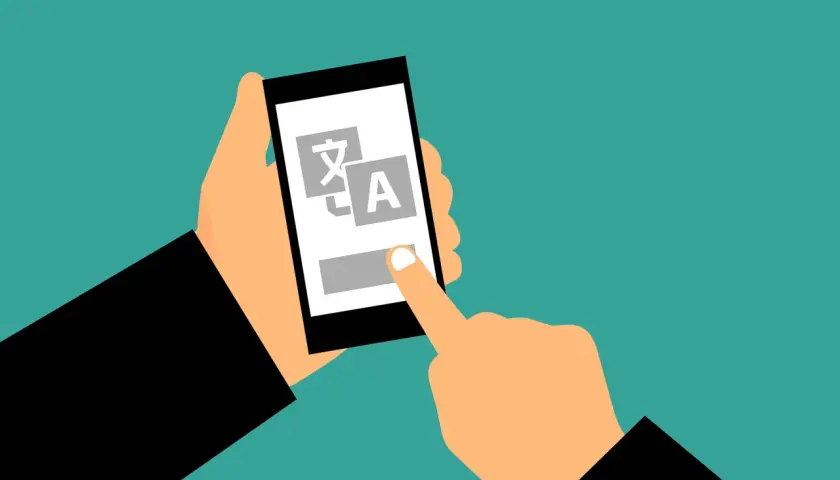 Dłoń trzyma smartfon z ikonami języków, a palec dotyka ekranu. Także czy tak że, tłumaczenie jest na wyciągnięcie ręki.