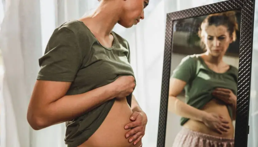 Mujer se mira al espejo, tocándose el abdomen con preocupación por la hinchazón después de comer.
