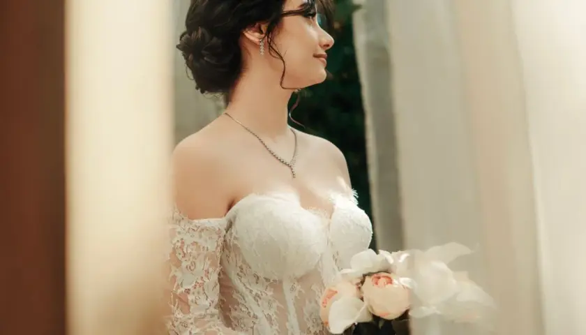 Eleganckie, najmodniejsze upięcia fryzury na wesele. Panna młoda w tiarze i sukni z koronki trzyma bukiet.