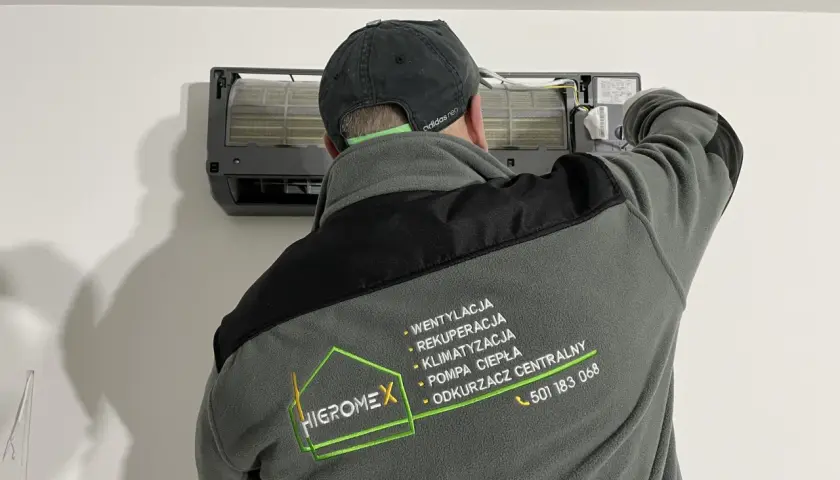 Technik w czapce z daszkiem montuje klimatyzator. Na kurtce logo firmy Higromex.