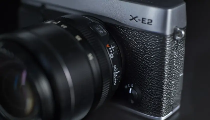 Srebrny aparat Fujifilm X-E2 z obiektywem. Widoczny napis "X-E2" i pokrętło wyboru trybu.