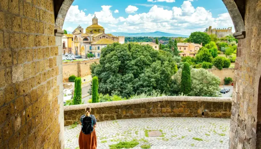 A traveler pauses, backpack ready, gazing at the historic Italian landscape. This view, perhaps on a journey from Viterbo to Rome, showcases ancient architecture and lush greenery under a bright sky.