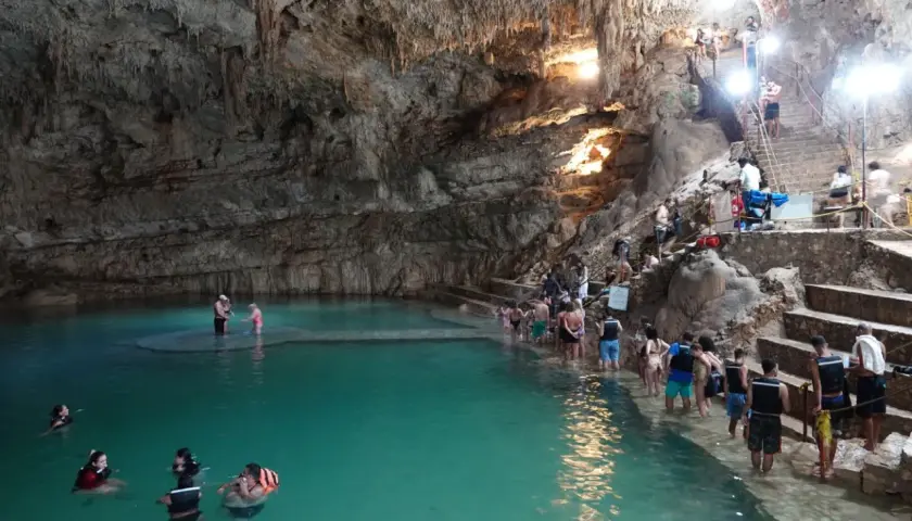 Menschen schwimmen und stehen im türkisfarbenen Wasser einer Höhle in Valladolid, Mexiko. Treppen führen zu einer Plattform, auf der sich weitere Besucher aufhalten.