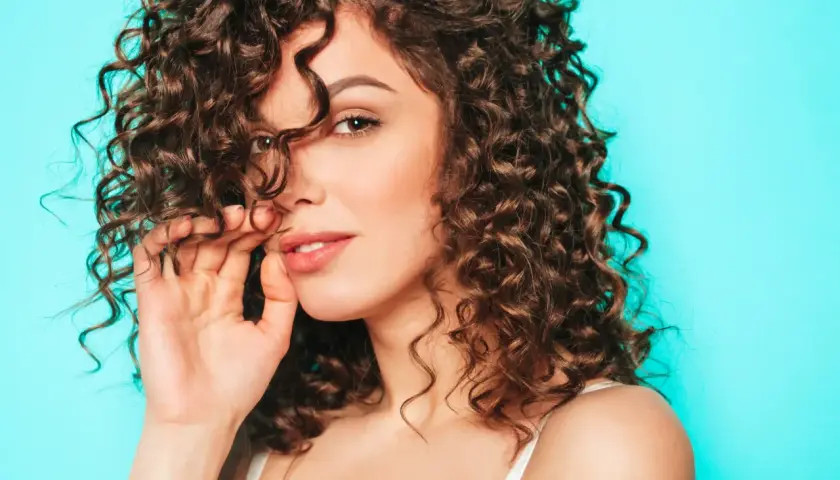 Mujer con cabello castaño y rizos definidos, tocando su cabello con la mano, sobre un fondo azul turquesa.