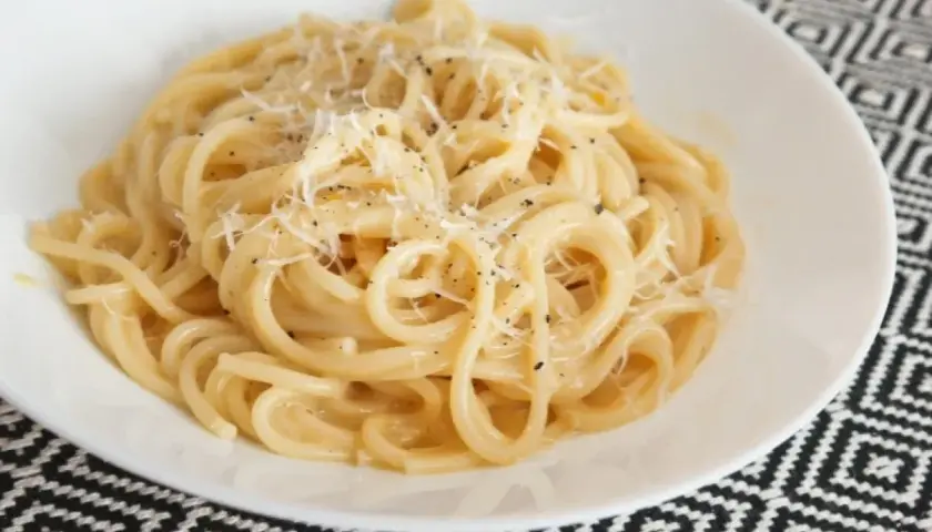 Pyszne spaghetti carbonare, posypane startym parmezanem i świeżo mielonym pieprzem, w białej misce na wzorzystym obrusie.