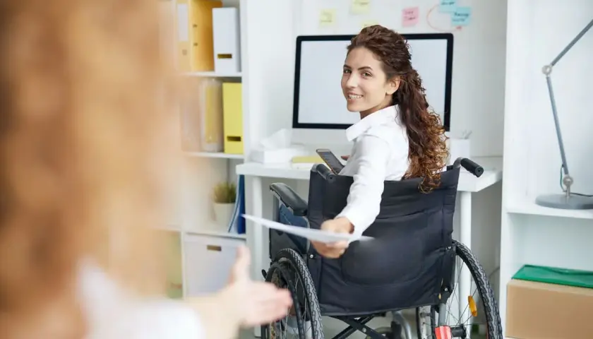 Donna in carrozzina sorride, porgendo documenti. La legge 68/99 offre vantaggi per il lavoratore, promuovendo inclusione e pari opportunità.