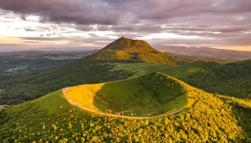 Vue panoramique du Puy de Dôme au coucher du soleil, avec un chemin serpentant sur une colline verdoyante. Idéal pour un séjour en camping-car.