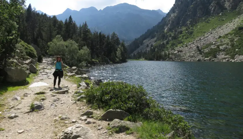 Una mujer camina por un sendero rocoso junto a un estany llong, rodeado de montañas y pinos.