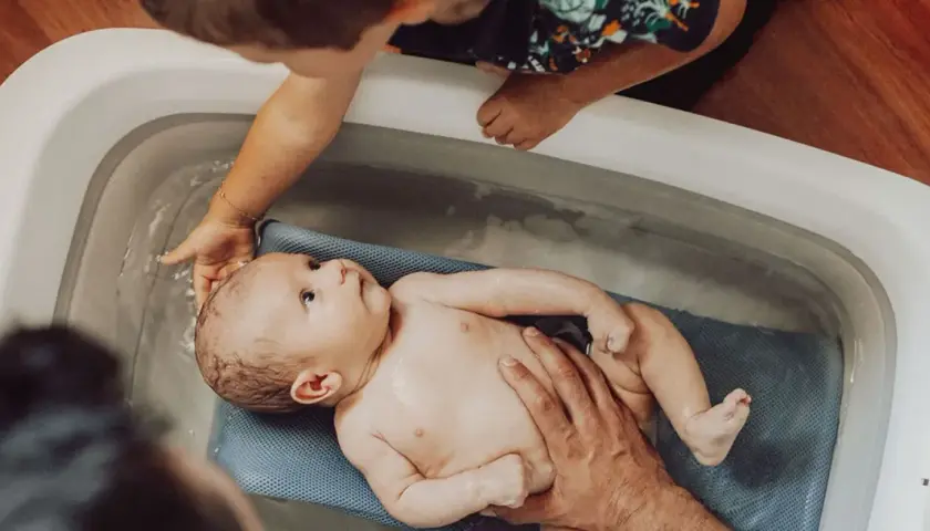Delikatna kąpiel noworodka w wanience. Starsze dziecko z zaciekawieniem obserwuje maluszka.