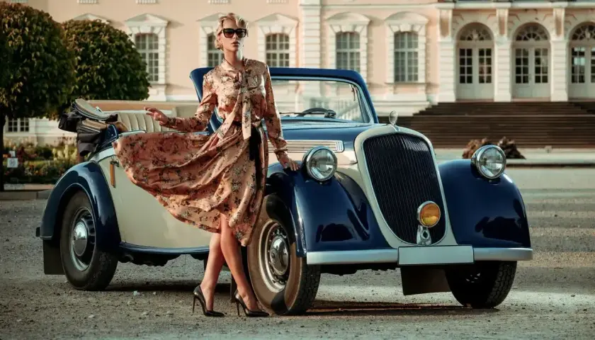 Elegancka kobieta w kwiecistej sukience i okularach przeciwsłonecznych opiera się o zabytkowy samochód. Stylowe lata 20. w pełnej krasie.
