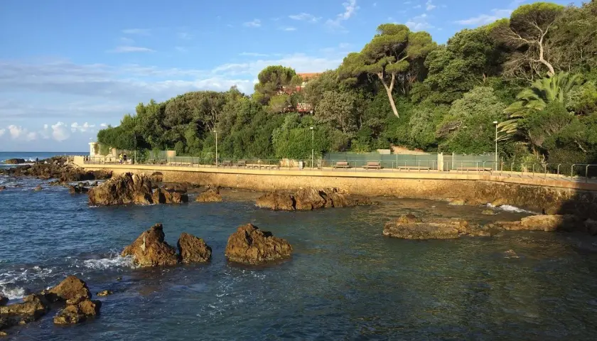 Promenade en bord de mer à Castiglioncello, Italie, avec des rochers dans l'eau et une colline verdoyante.