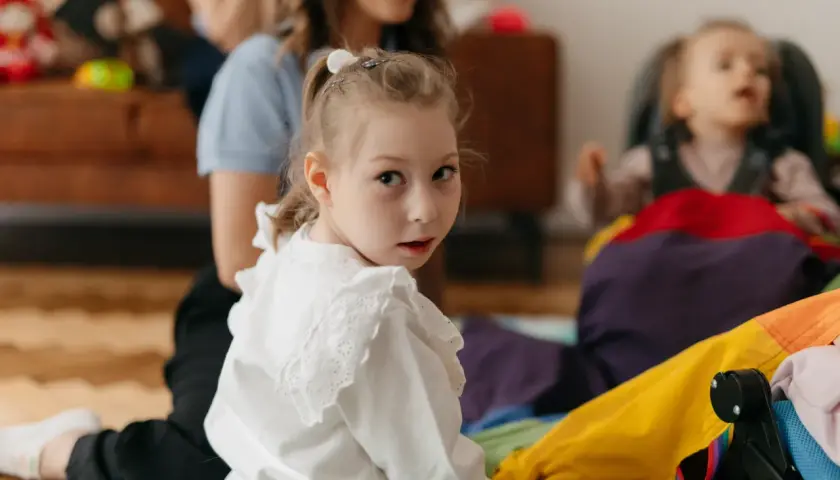 Dziewczynka w białej bluzce patrzy w obiektyw, w tle kobieta i dziecko w wózku.
