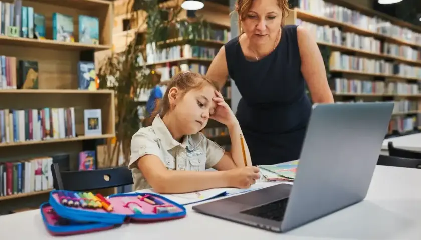 Dziewczynka z ołówkiem w dłoni, zamyślona nad zadaniem, przygotowuje podanie do szkoły średniej. Nauczycielka wspiera ją w bibliotece.