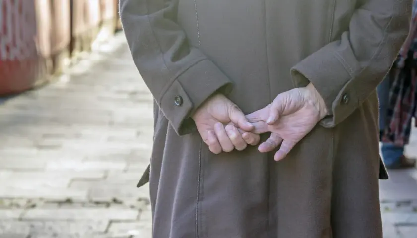 Osoba w brązowym płaszczu, z rękami splecionymi za plecami, stoi na chodniku.