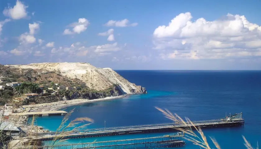 Panoramica delle isole Lipari con un porto e moli che si estendono nel mare blu intenso sotto un cielo azzurro con nuvole bianche.