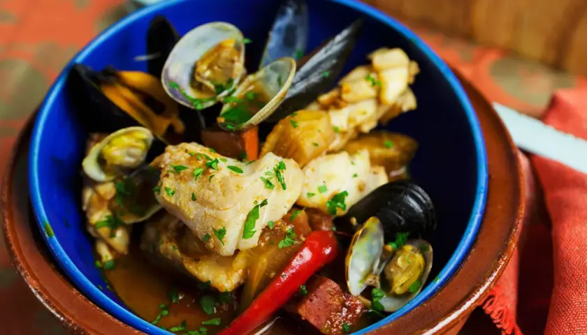 Delicioso bacalao con almejas y gambas, servido en un cuenco azul con perejil picado y un chile rojo.
