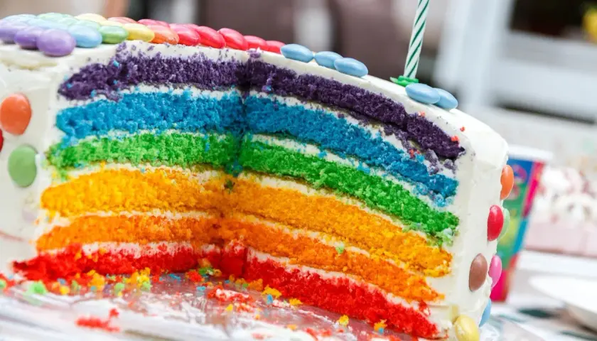 Kolorowy tort tęczowy z warstwami ciasta w odcieniach czerwieni, pomarańczu, żółci, zieleni, błękitu i fioletu, ozdobiony cukierkami.
