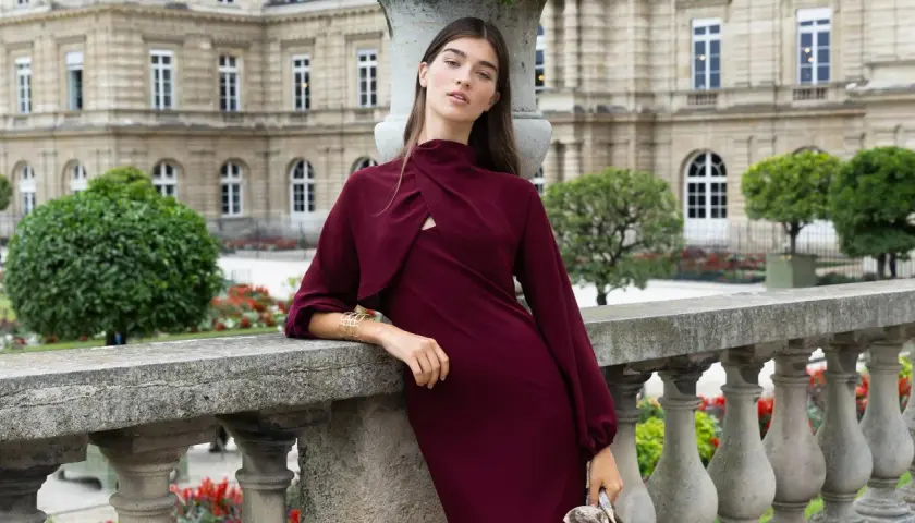 Mujer con un elegante vestido granate, apoyada en una barandilla de piedra, con un edificio histórico y jardines de fondo.