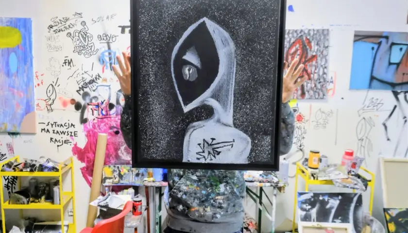 Osoba trzyma obraz z postacią w kapturze, przypominającą styl tatuażu graffiti. W tle ściany pokryte rysunkami i farbami.