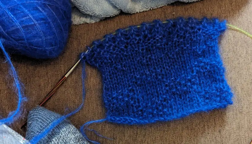 Un échantillon de tricot bleu vif, avec une pelote de laine assortie et une aiguille. La jauge maille est bien visible sur le tissu.