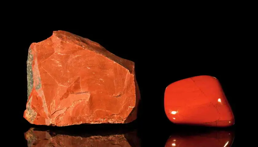 Zwei rote Jaspis-Arten: ein roher Stein und ein polierter Trommelstein, die auf schwarzem Hintergrund reflektieren.