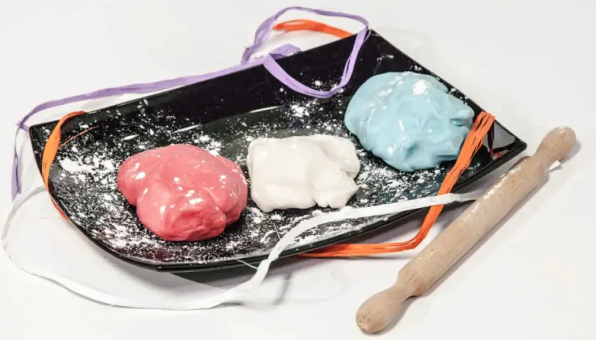 Tre panetti di pasta di zucchero colorata (rosa, bianca, azzurra) su un piatto nero con nastri colorati e un mattarello.