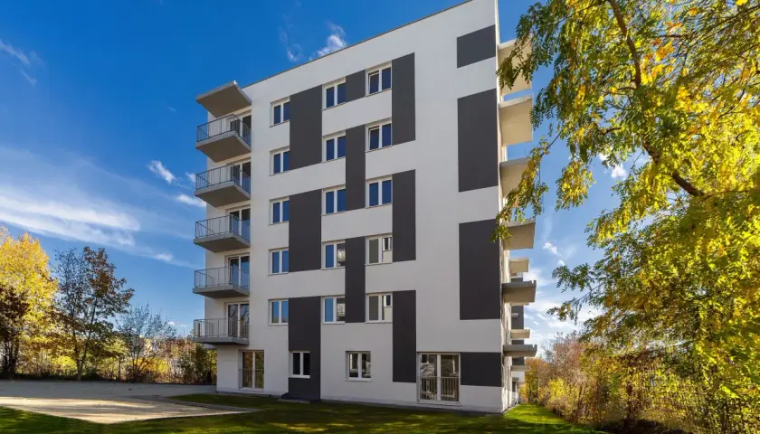 Nowoczesny budynek mieszkalny z balkonami i geometrycznym wzorem na elewacji, otoczony zielenią.