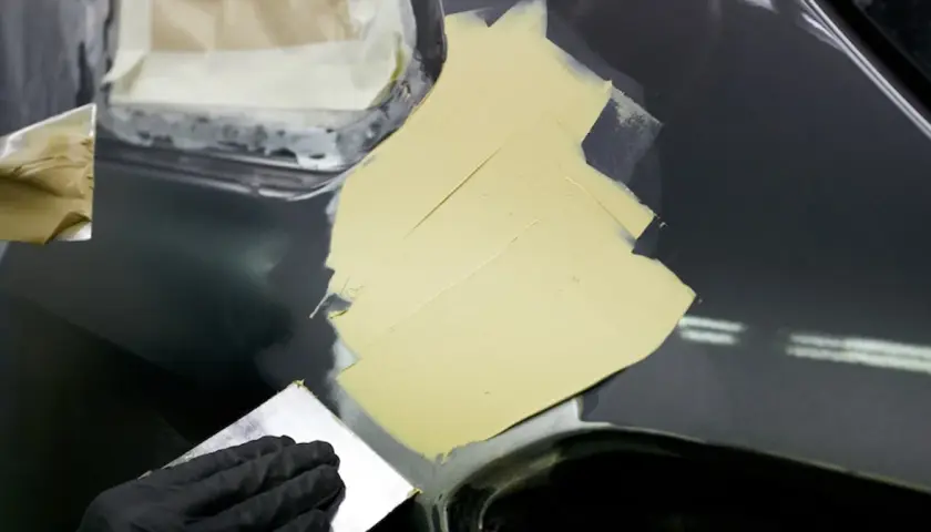 Ręka w czarnej rękawiczce rozprowadza szpachlę na karoserii, pokazując, jak szpachlować samochód.