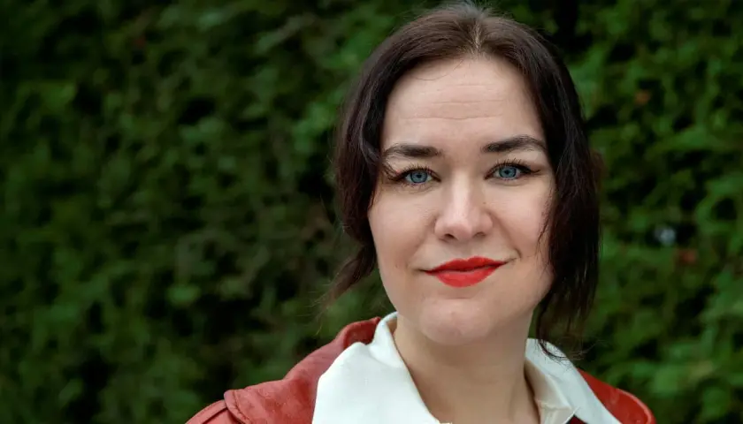 Camilla Sten med blå ögon och rött läppstift, klädd i en röd jacka, ler mot kameran.