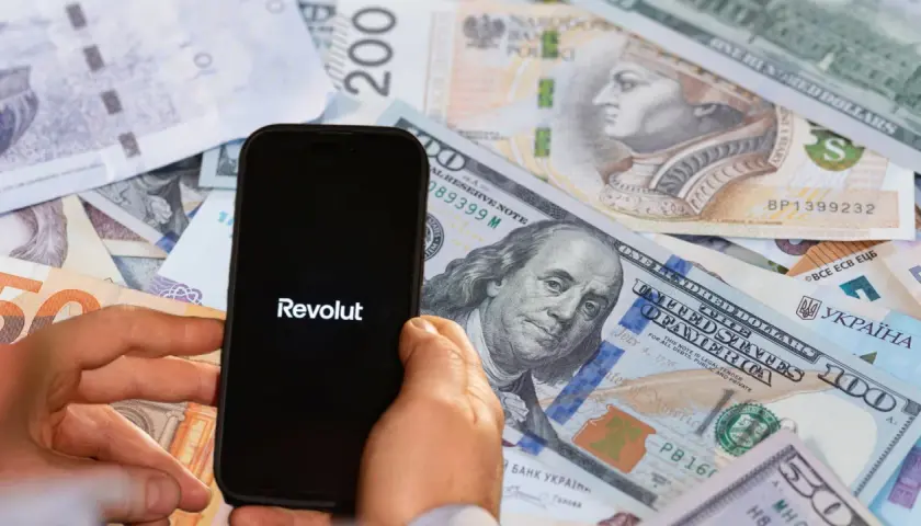 Telefon z logo Revolut na tle różnych walut. Może to symbolizować globalne finanse lub potencjalne zajęcie komornicze w kontekście międzynarodowych transakcji.